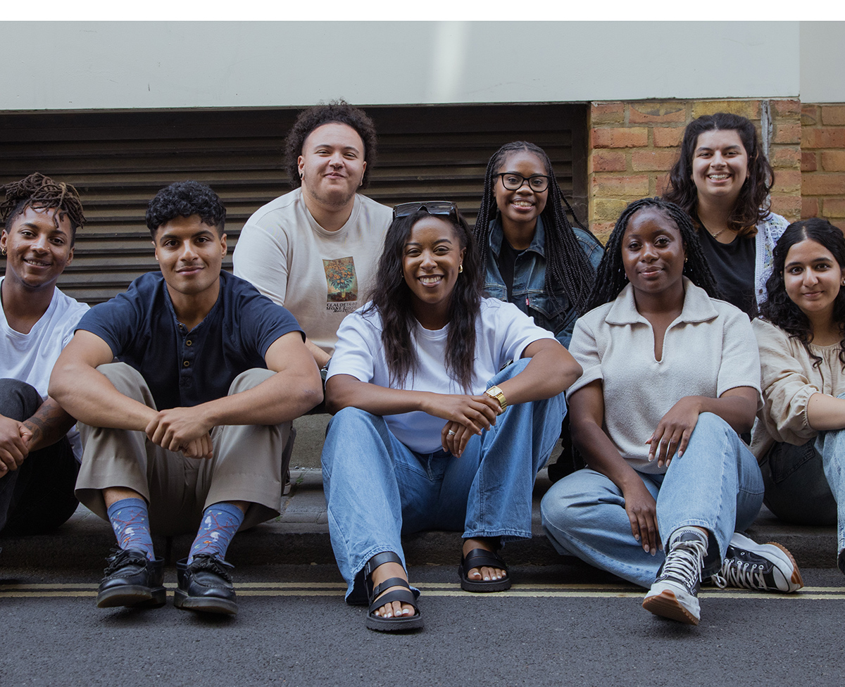 A diverse group of young people are sat on a street curb, looking directly at the camera and smiling.