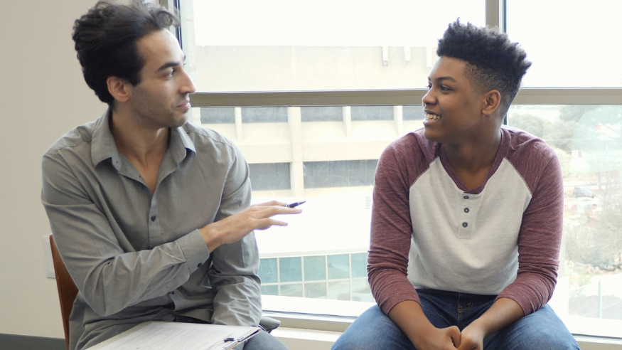 Male therapist sat on a chair, left of image, with a clipboard. He is gesticulating with his right hand whilst holding a pen. The teen boy is also sat on a chair, to the image right, looking at the therapist and smiling.