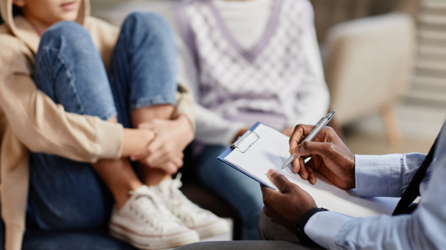 A therapy session. In the right foreground we seen an in-focus clipboard, with a therapist visibly taking notes. In the background and slightly out of focus, we seen a women sat on a chair with her feet also on the chair, and her arms wrapped around her legs. Another person is sat casually to her left. We can't see either of their faces.