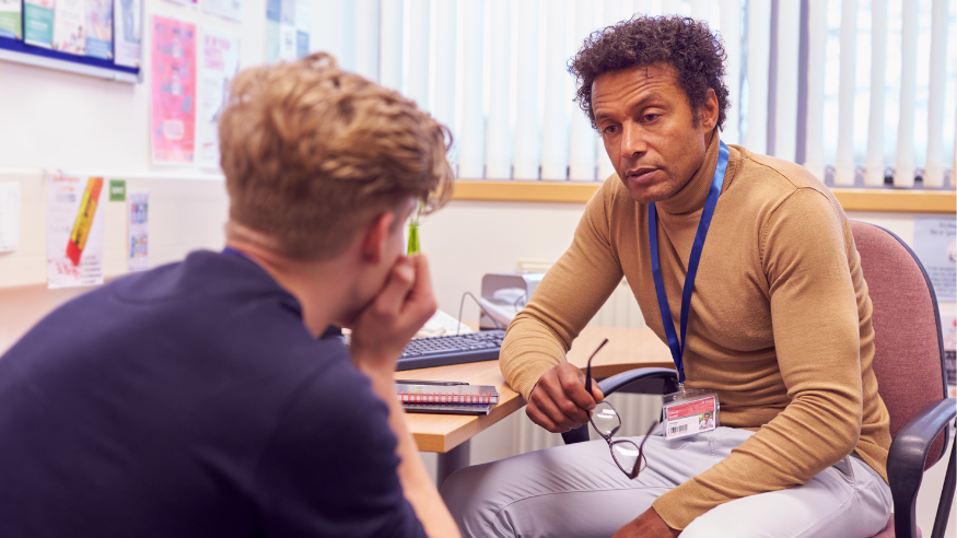Student in the foreground with back to camera, sat on a chair. His front of him, a councillor with a lanyard, looking thoughtfully at the young man.