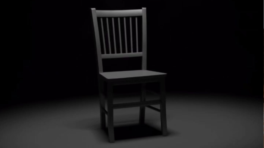 A grey chaired positioned at a slight angle, in a dark room. The chair is lit by an overhead spotlight.