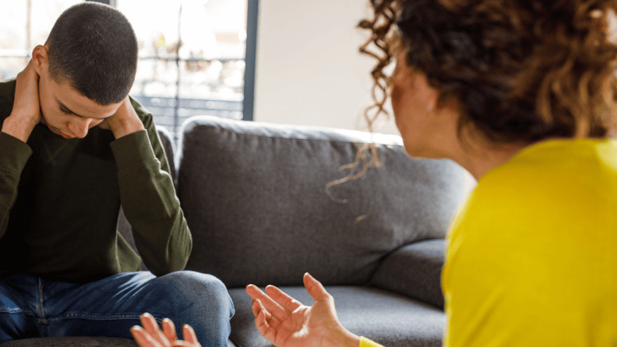 Female therapist talking with teenage boy. Therapist is position right of frame in the foreground. Teenage boy is left of frame, sat on a sofa with head positioned down, and his hands holding the back of his neck, appearing uncomfortable.