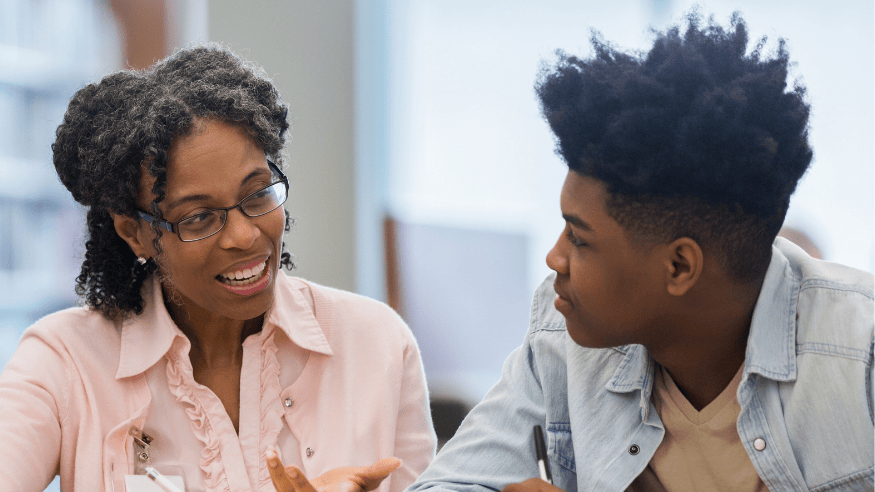 A women with glasses and a smart pink shirt is in conversation is with a teenage boy. They are sat down, towards the camera, facing one another. The mood is collaborative.