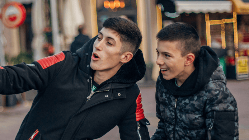 Two teenage boys filming themselves. The background is out of focus. They are both wearing puffer jackets.