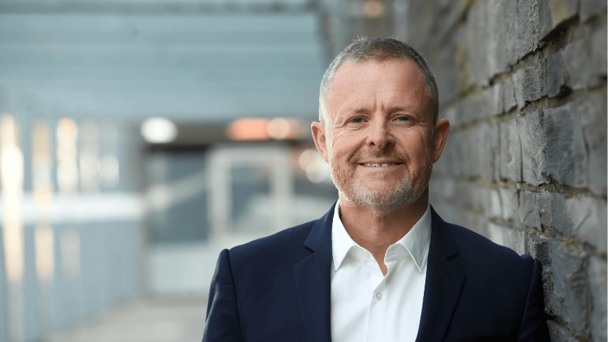 A three quarter head shot of a man in a shirt and blazer. He has short grey hair, a slight smile and stubble. His left shoulder is up against a grey brick wall.