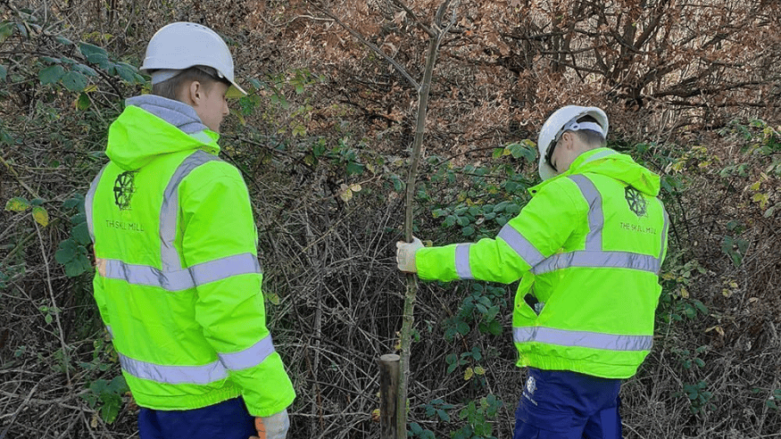 Two young men stood with their back to the camera. They are wear high-vis jacket with the words 'The Skill Mill' emblazoned across the back. They are wearing hard hats. The young man to the right has his left had on a young tree. They are both focused on the tree- and appear to be inspecting it.
