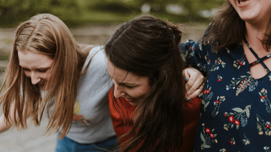 Three women with their arms over one-another's shoulders, laugh.