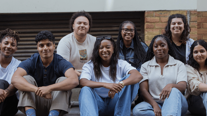 A group of young people, looking at the camera smiling. They are sat in a row of two. They are on a London street.