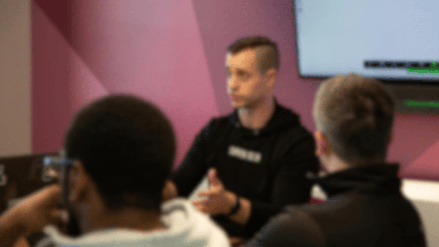 A blur photo a man in a black jumper speaking in a discussion, whereby attendees appear sat on chairs in a circle. Their are men in the foreground with their backs to the camera.