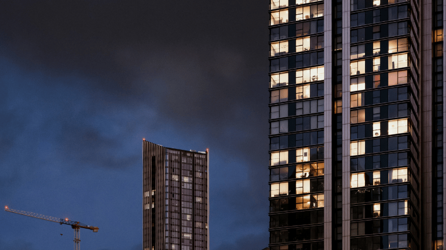 A tower block to the right. A splattering of lights are on. In some windows we can see furnishings, and the occasional person. In the middle of the shot, another tower, further away, so we can see he top of it. To the bottom left, a crane. It is dusk, and the light is low.